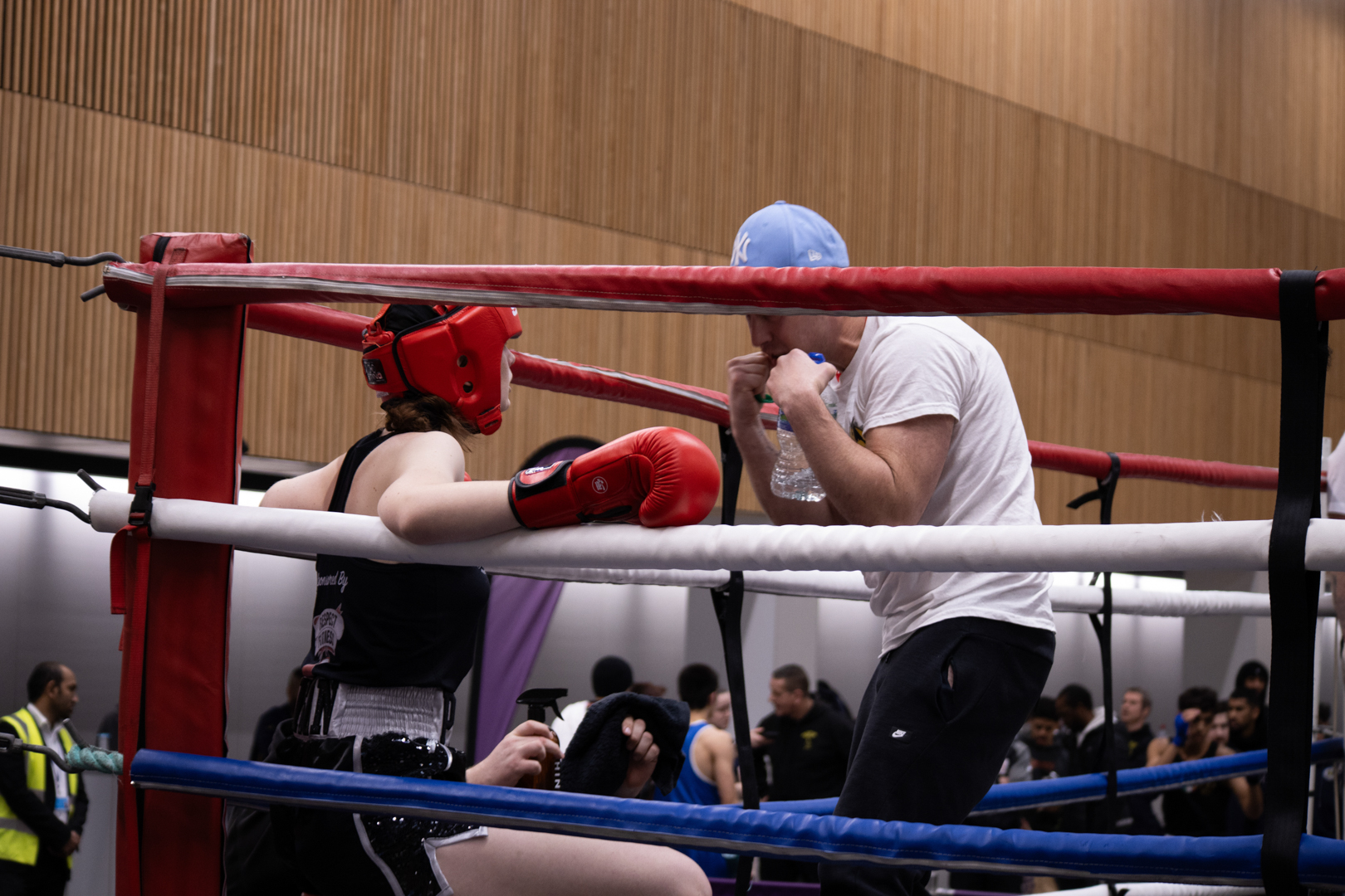 A boxer in red gear sits in a corner of a boxing ring while a coach or trainer speaks to them.