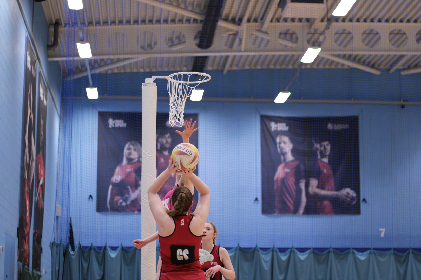 Netball player in a shoots while being defended in an indoor gym.