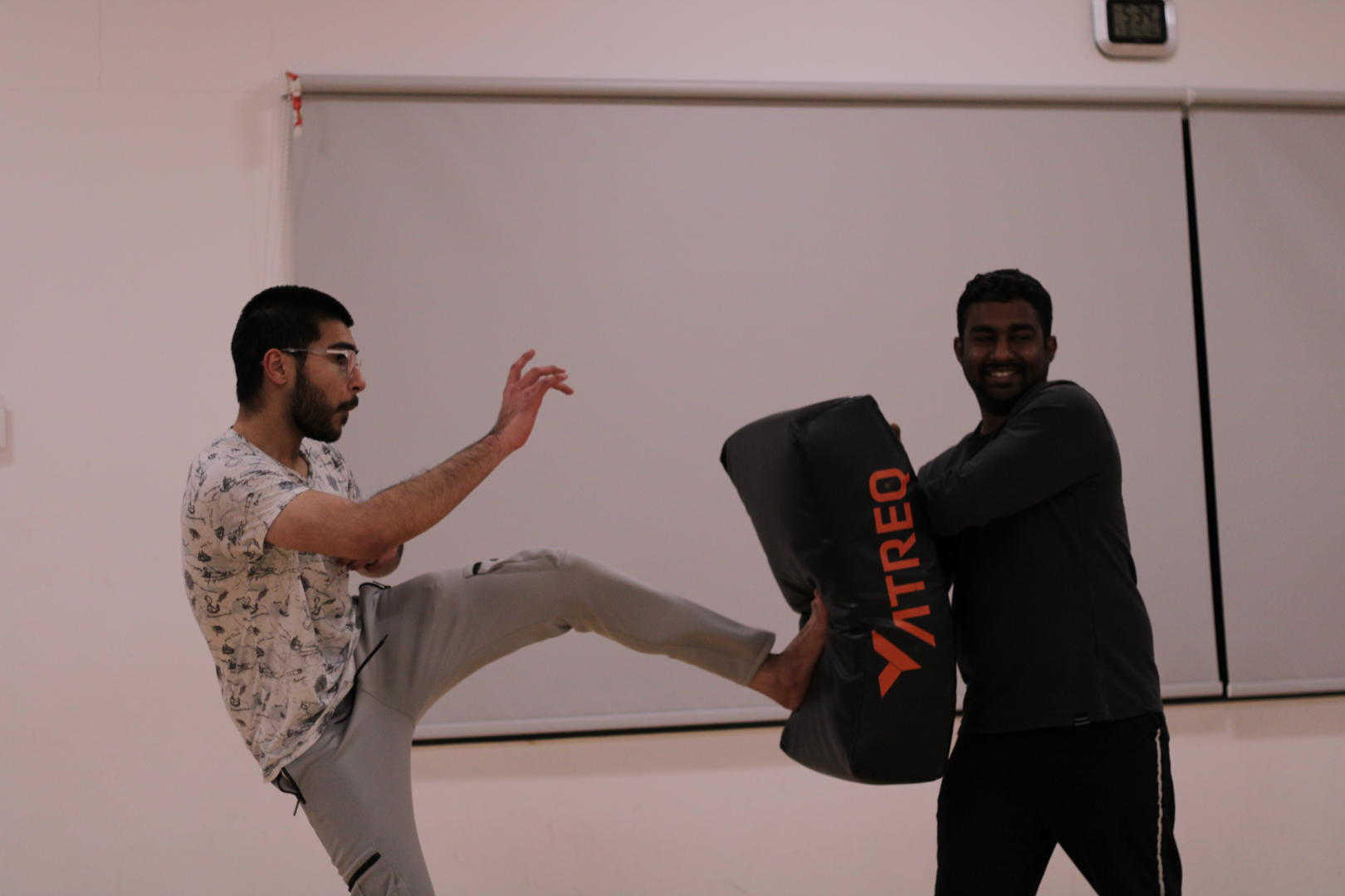 One person kicks a padded shield held by another person in a martial arts or self-defense practice.