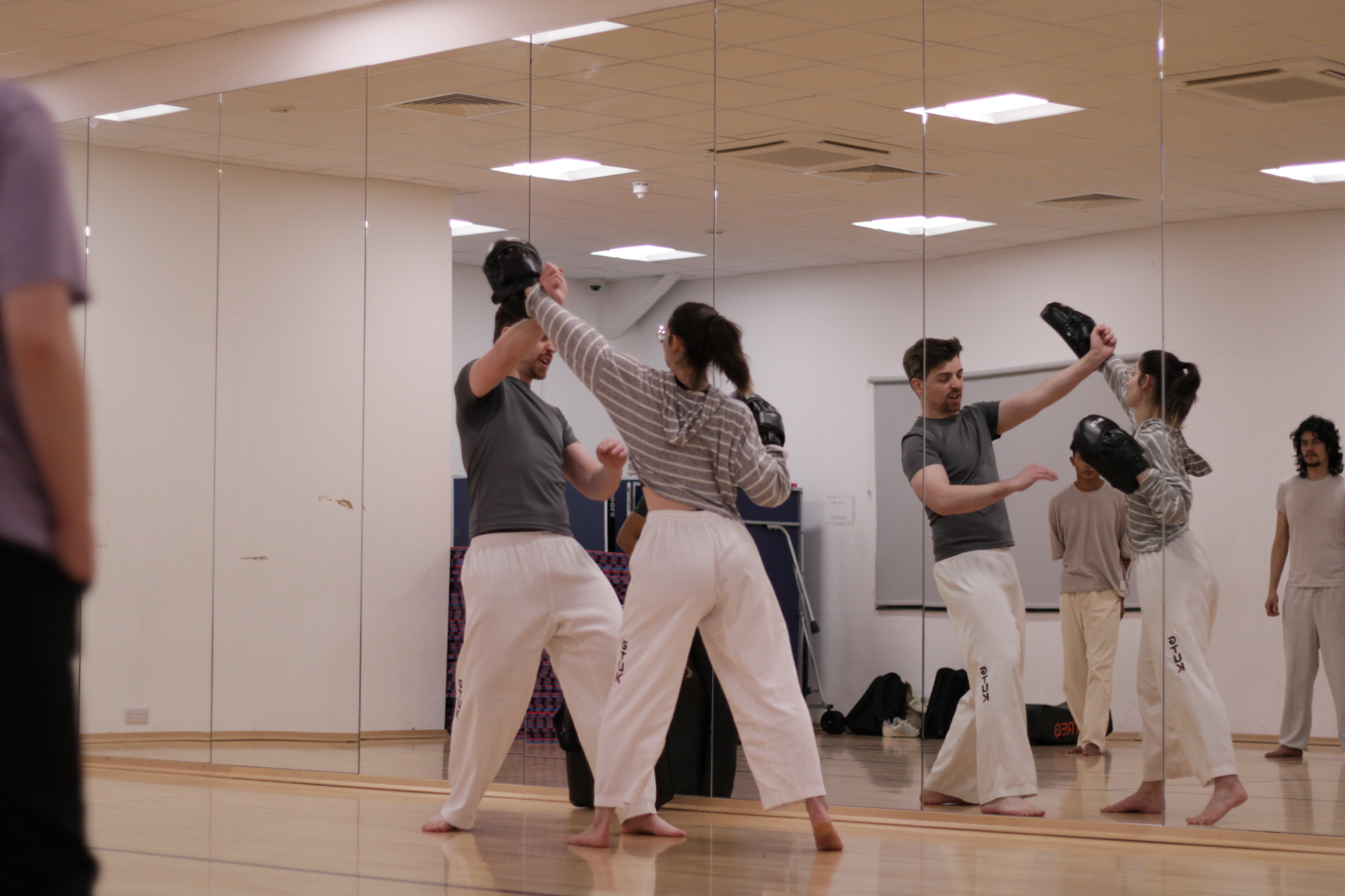 People practicing martial arts in a mirrored studio, wearing uniforms and performing paired exercises.