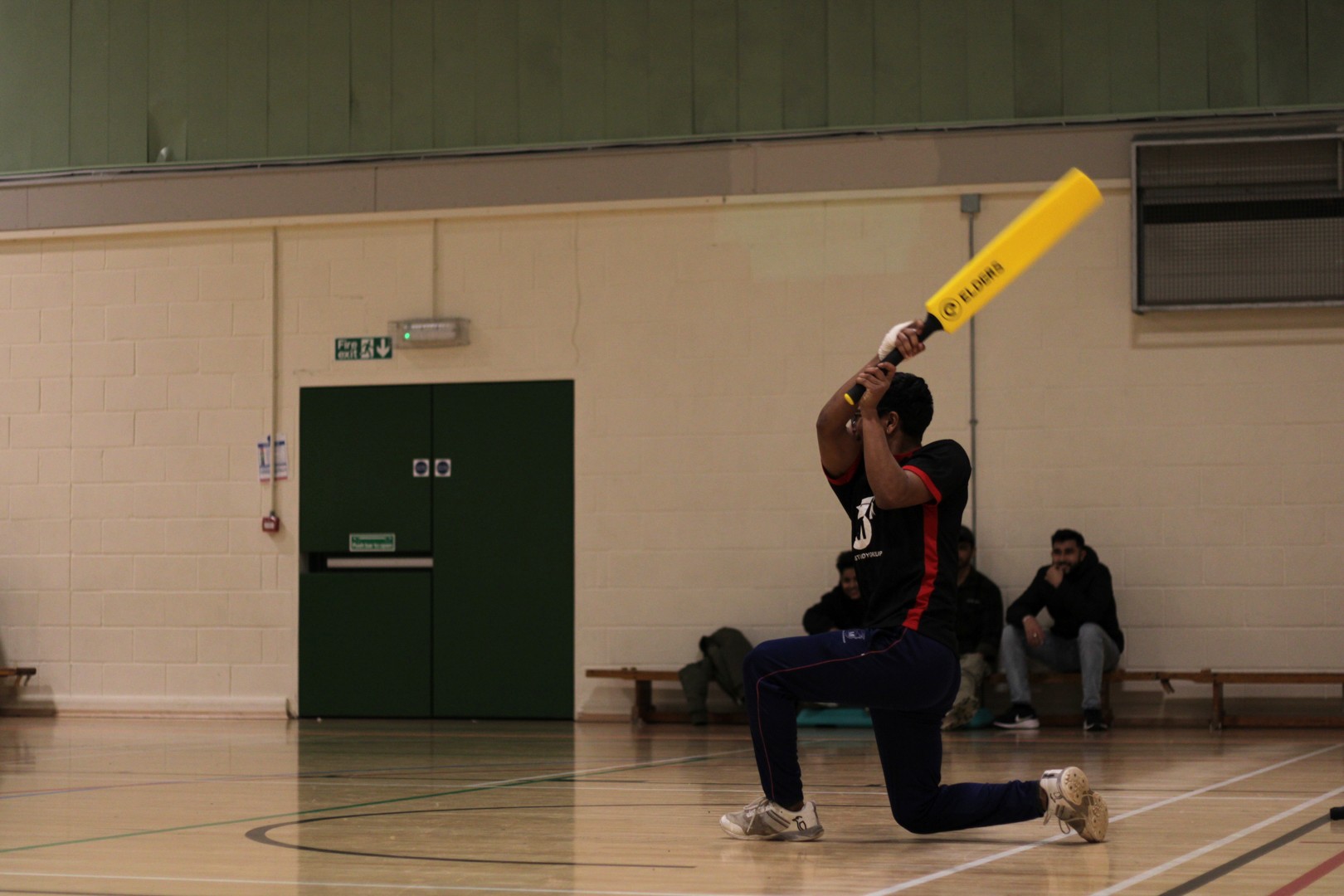Person kneeling indoors swings a yellow plastic cricket bat, with others seated against the wall in the background.