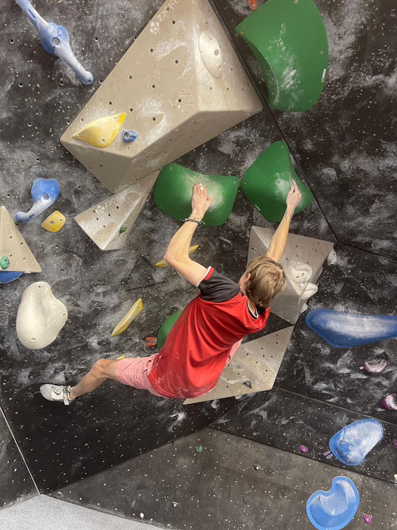 Student in a red shirt climbing an indoor bouldering wall, reaching for green holds.