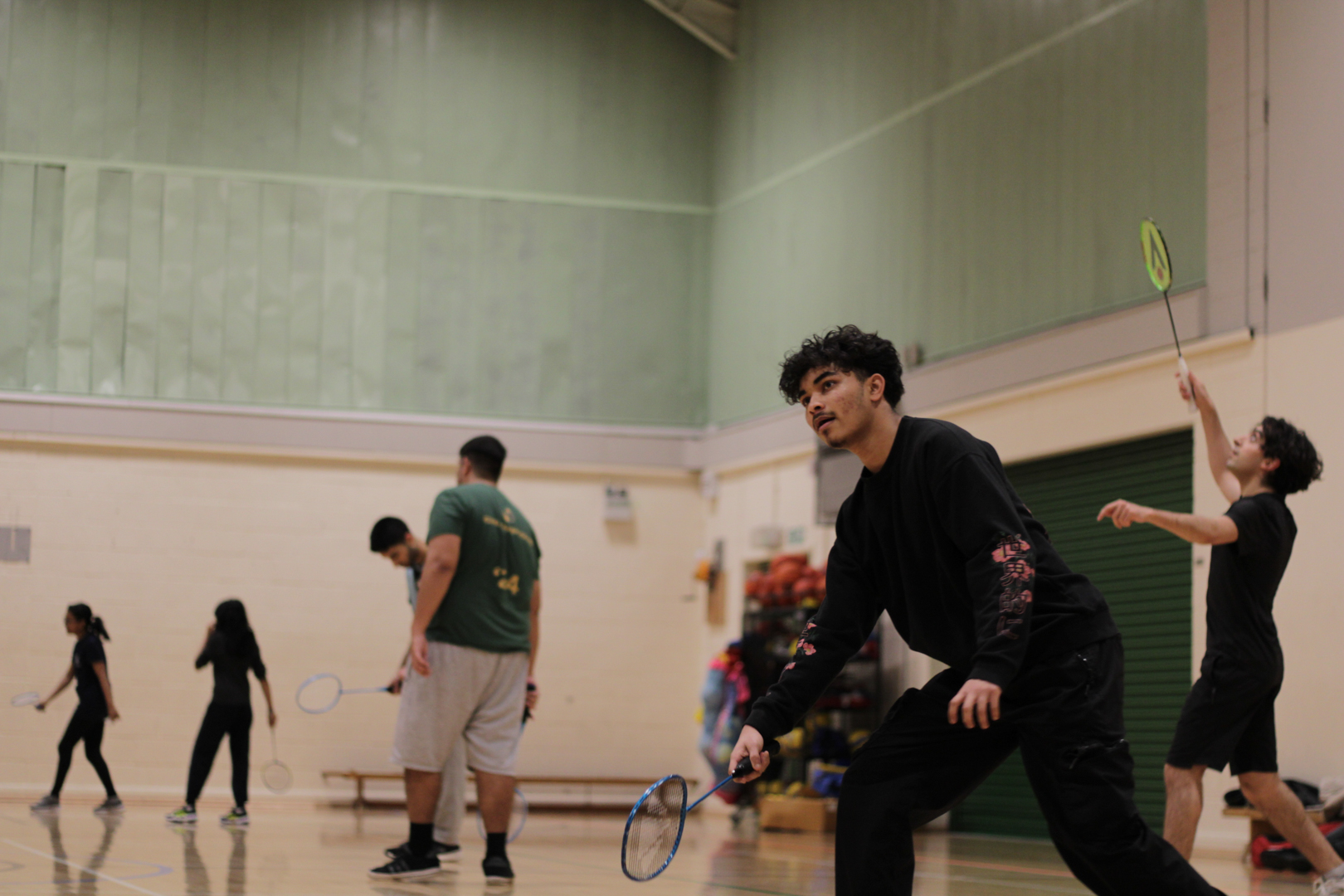 People playing Badminton in an indoor gym, with one person in the foreground and others positioned around the court.