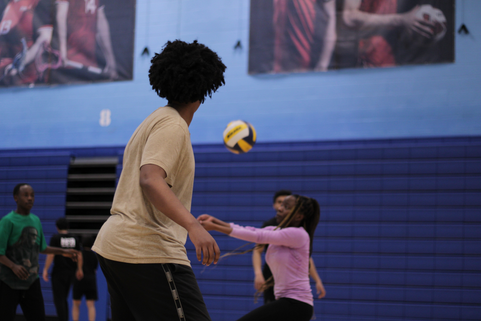 Two people in a gym playing with a yellow and blue volleyball, with others watching in the background.