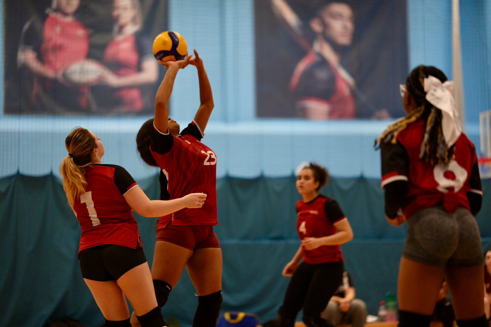 Women’s volleyball players in red and black uniforms jump and reach for a ball during an indoor match.