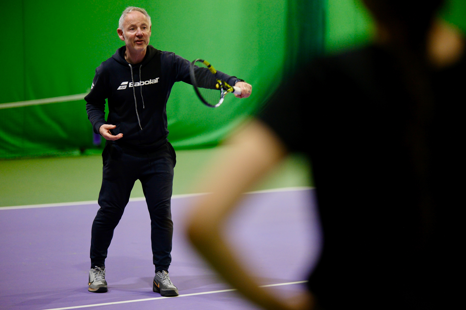 Tennis coach instructing a player on an indoor court with a green background.