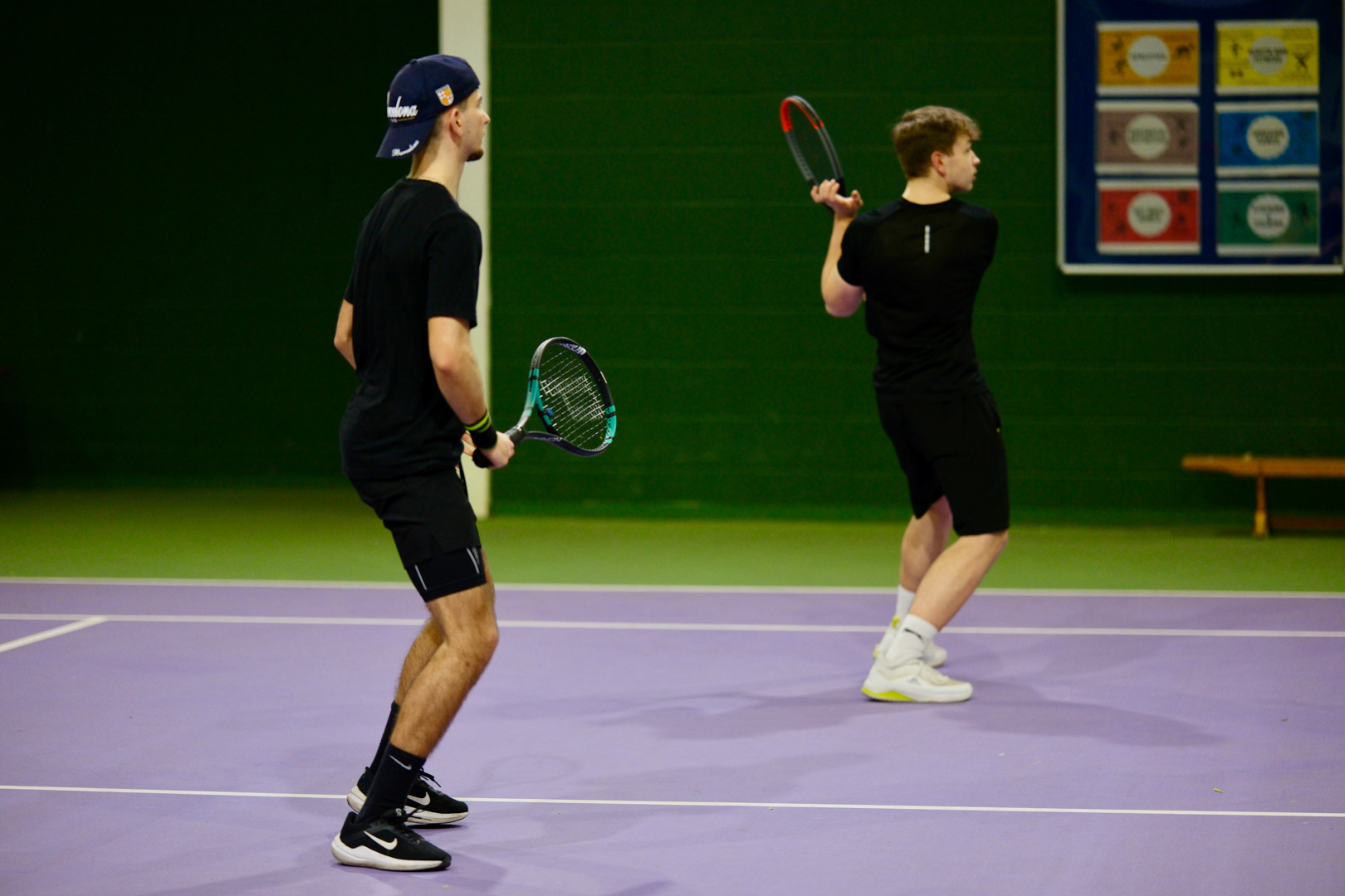 Two people playing tennis indoors on a purple court, both wearing black athletic clothing.