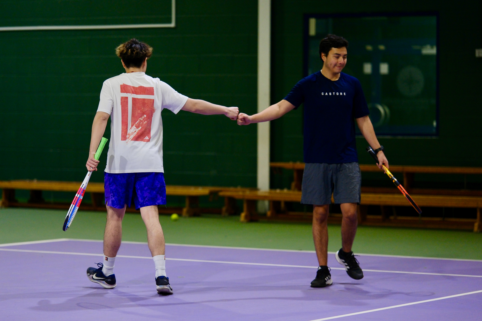 Two men on a tennis court give each other a fist bump while holding tennis rackets.