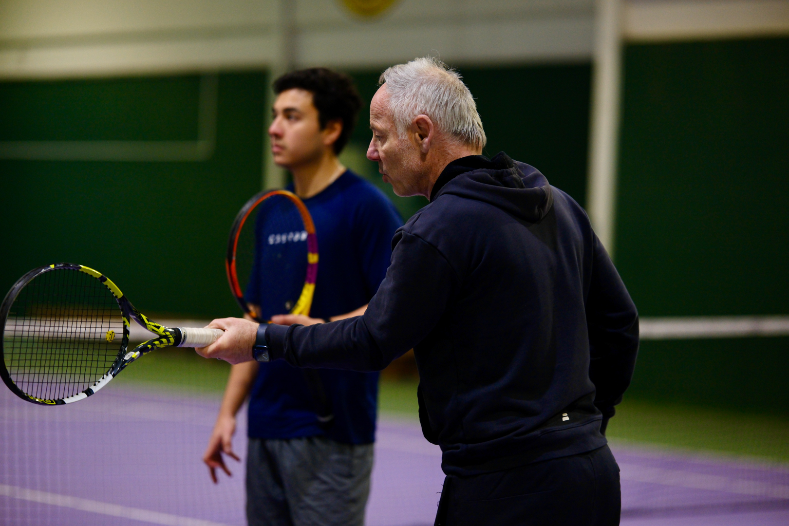 Older man instructing a younger man on a tennis court, both holding tennis rackets.