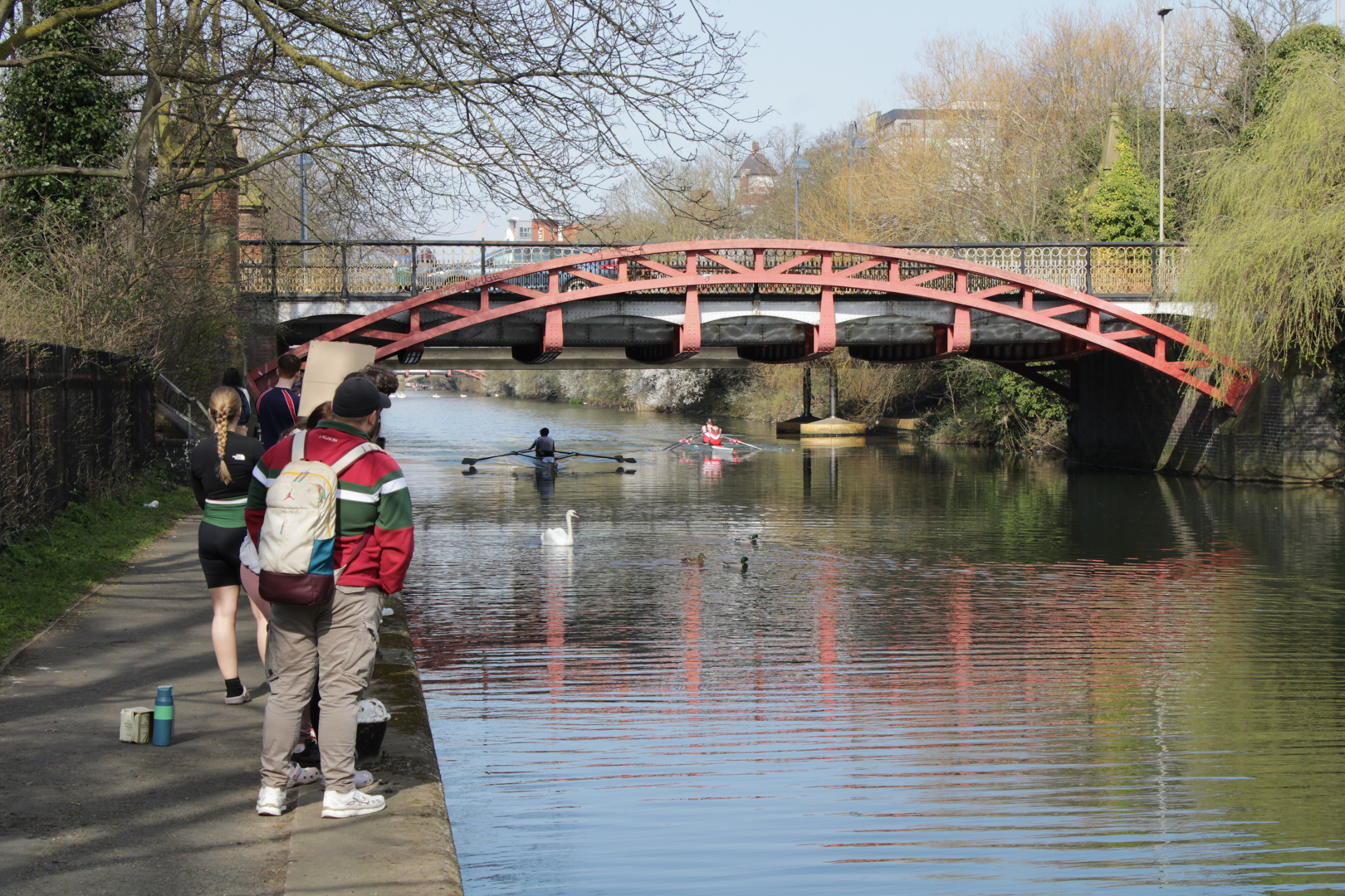 People walking along a canal path with people rowing past a bridge.