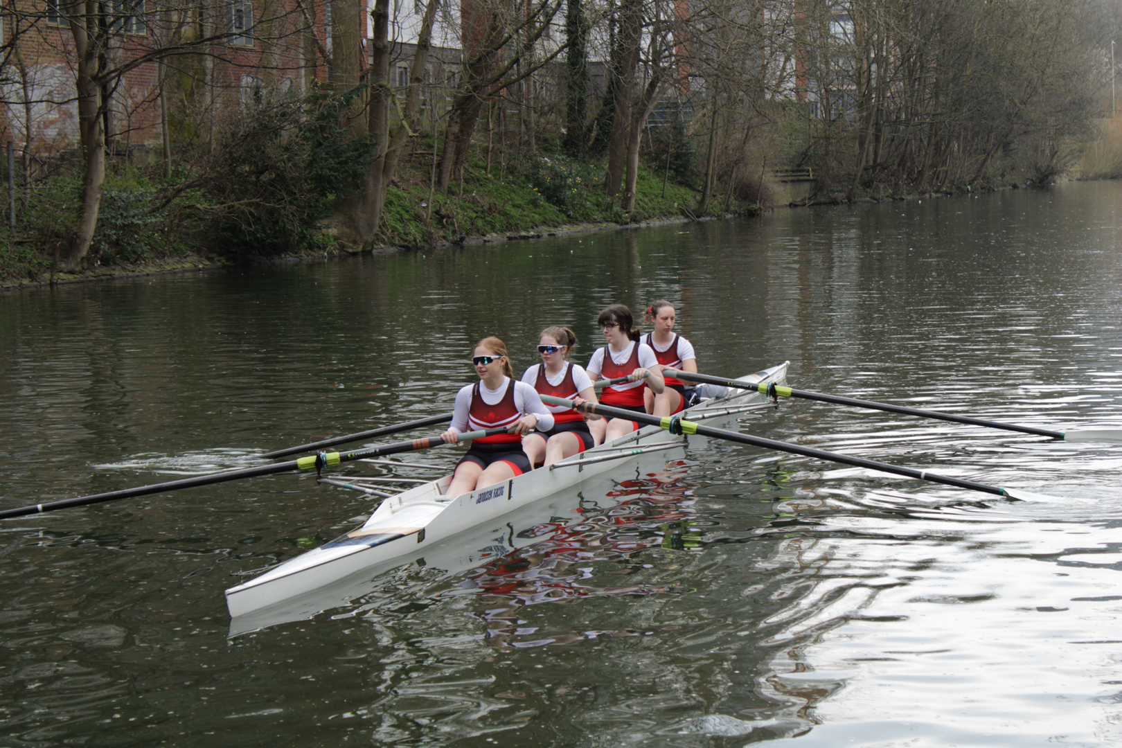 Four rowers in matching uniforms rowing a boat on a calm river with trees in the background.