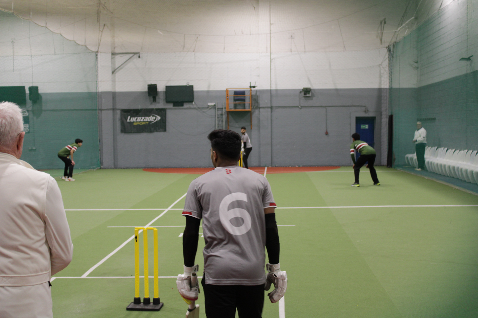 Indoor cricket game in progress, with players positioned on a green court and one player preparing to bat.