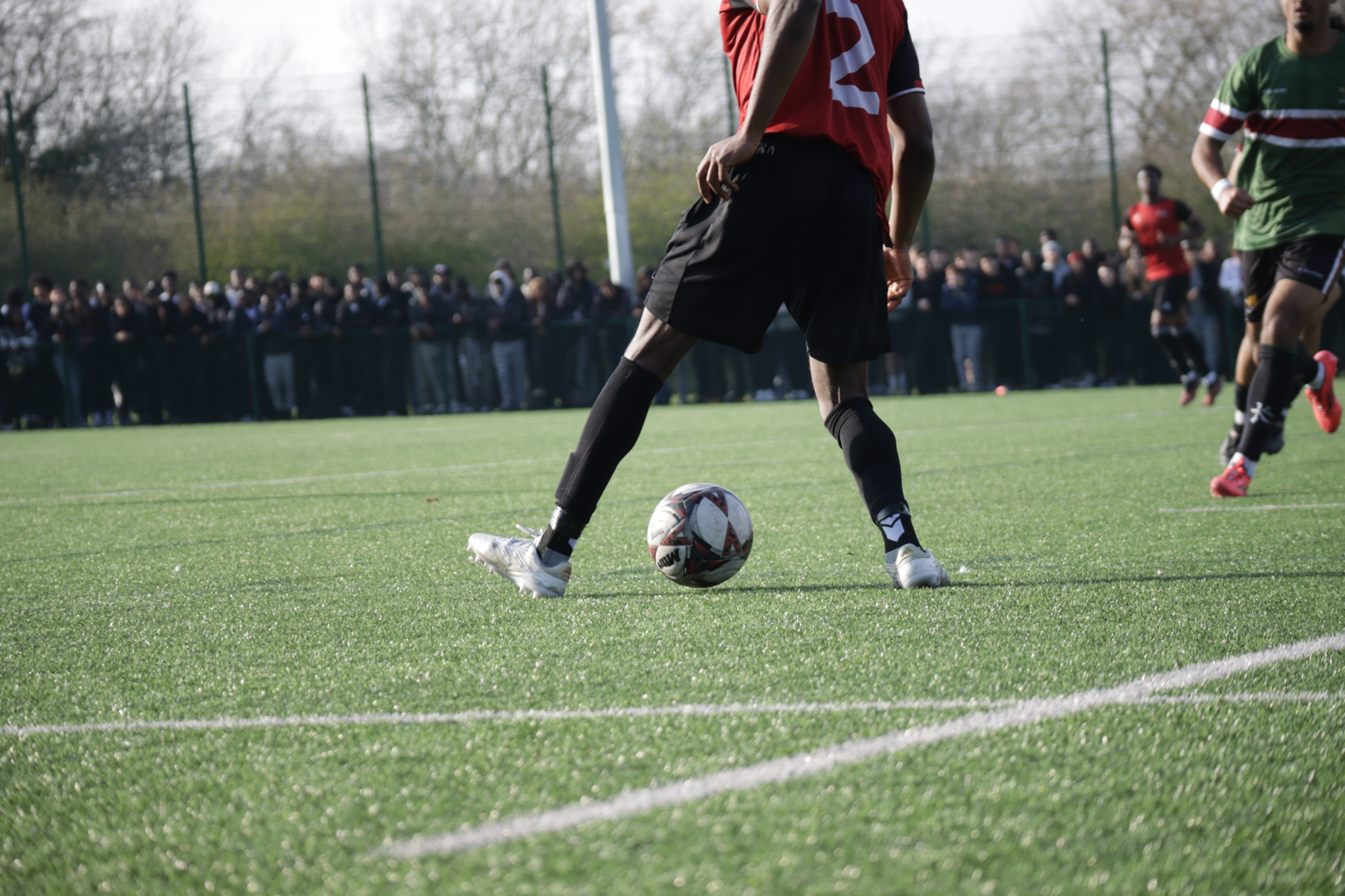 Football player in black shorts and red shirt dribbling a ball on a pitch during a match.