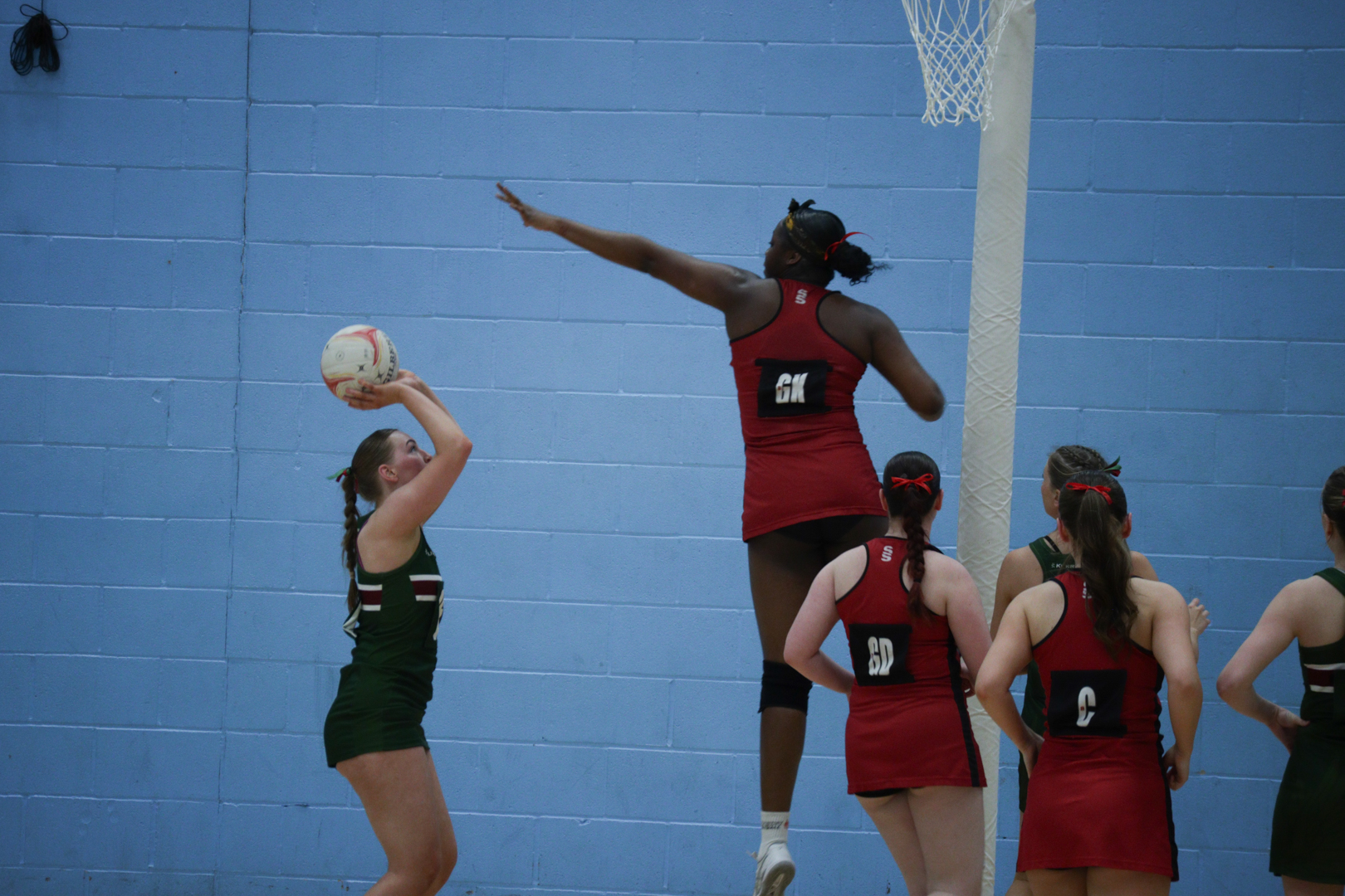 Players in a netball game, one in green preparing to shoot while a player in red jumps to block the shot.