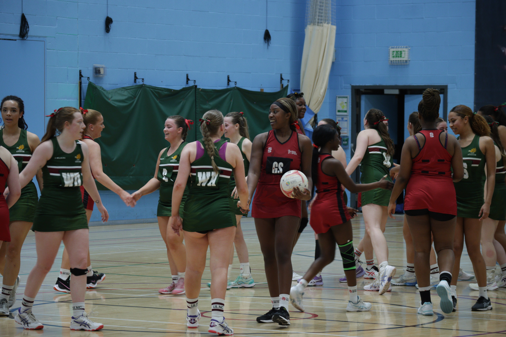 Two netball teams, one in green and one in red, shake hands on an indoor court.
