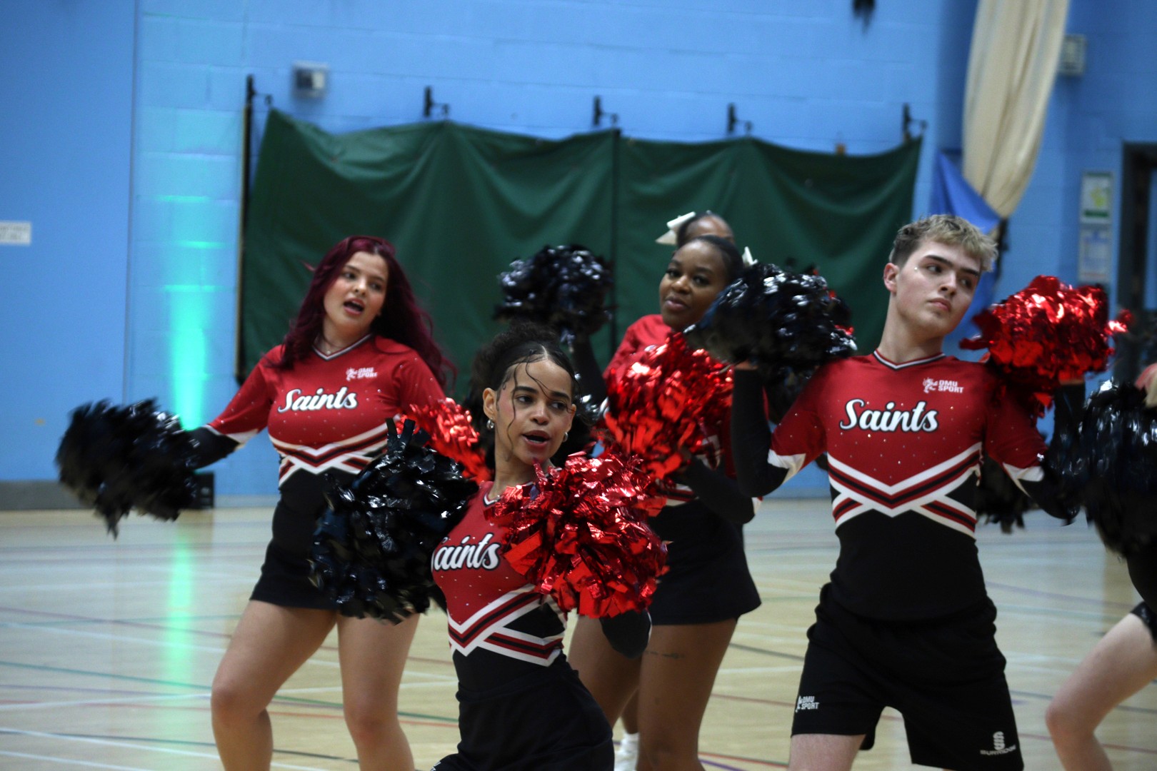 Four cheerleaders in red and black uniforms performing indoors with pom-poms raised.
