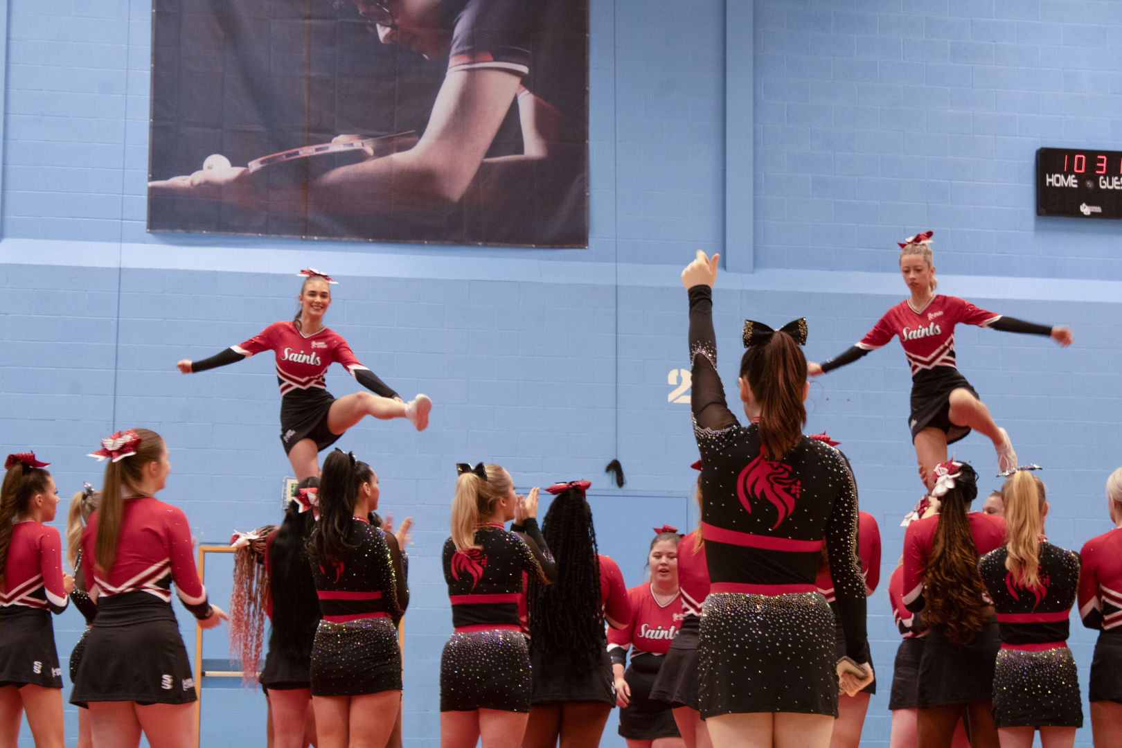 Cheerleaders in red and black uniforms perform stunts in a gymnasium with a blue wall and sports posters.