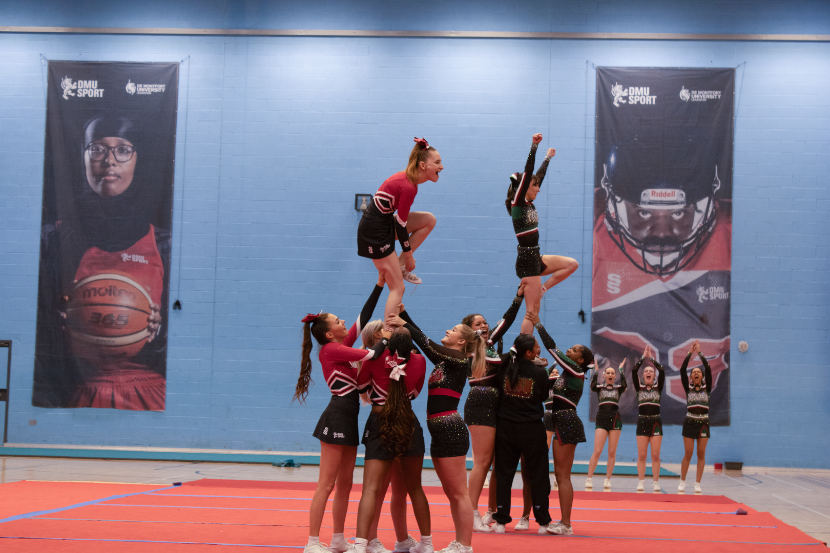 Cheerleading team performs a stunt routine indoors, with flyers lifted high above the bases on a gym mat.