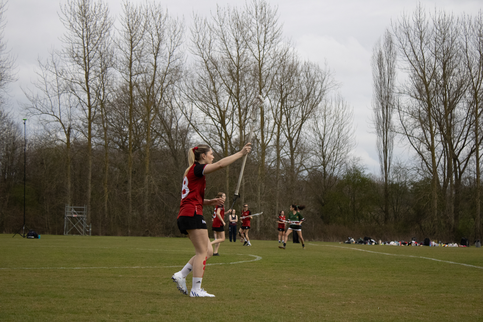 Lacrosse players in red and black kits on a grassy field with trees in the background.