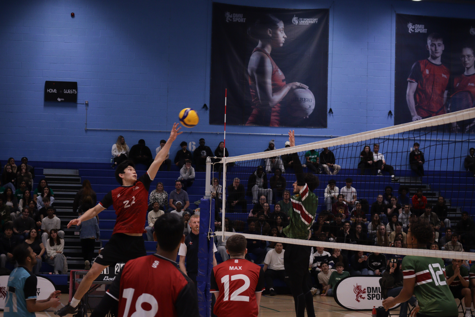 Two volleyball teams compete indoors, with one player jumping to spike the ball over the net as spectators watch.