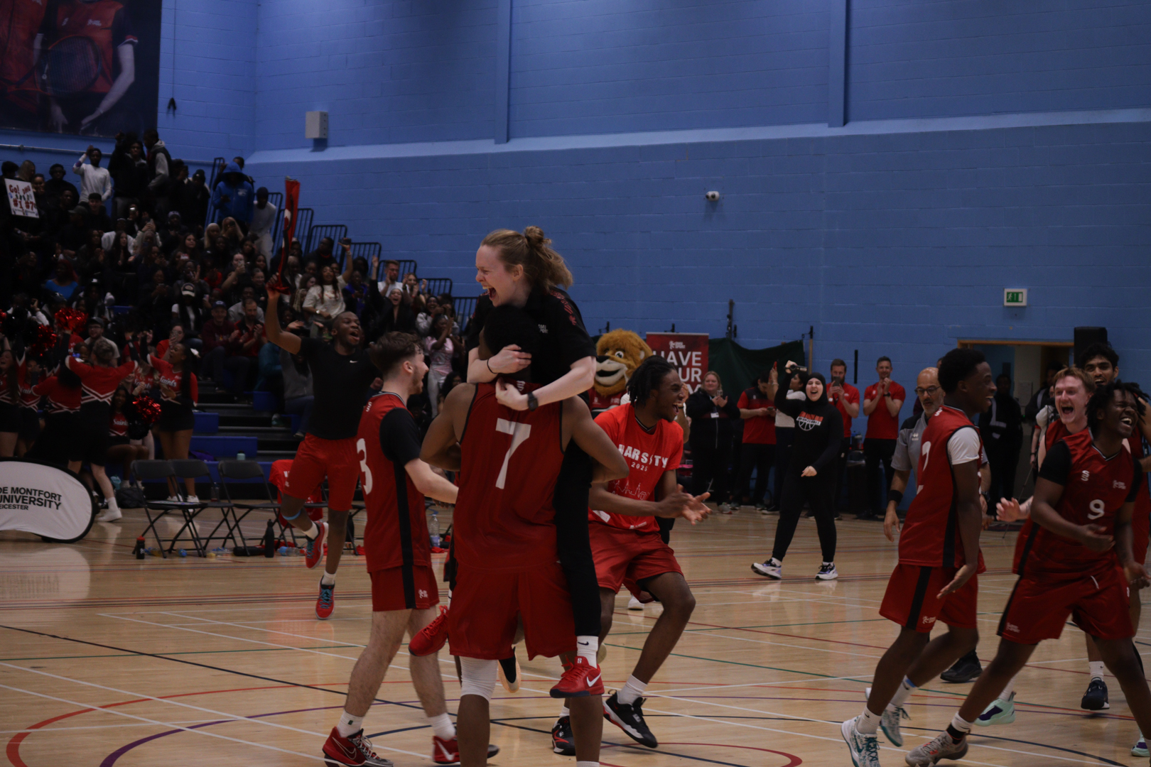 Basketball players in red kit celebrate on the court, lifting a coach, with spectators in the background.