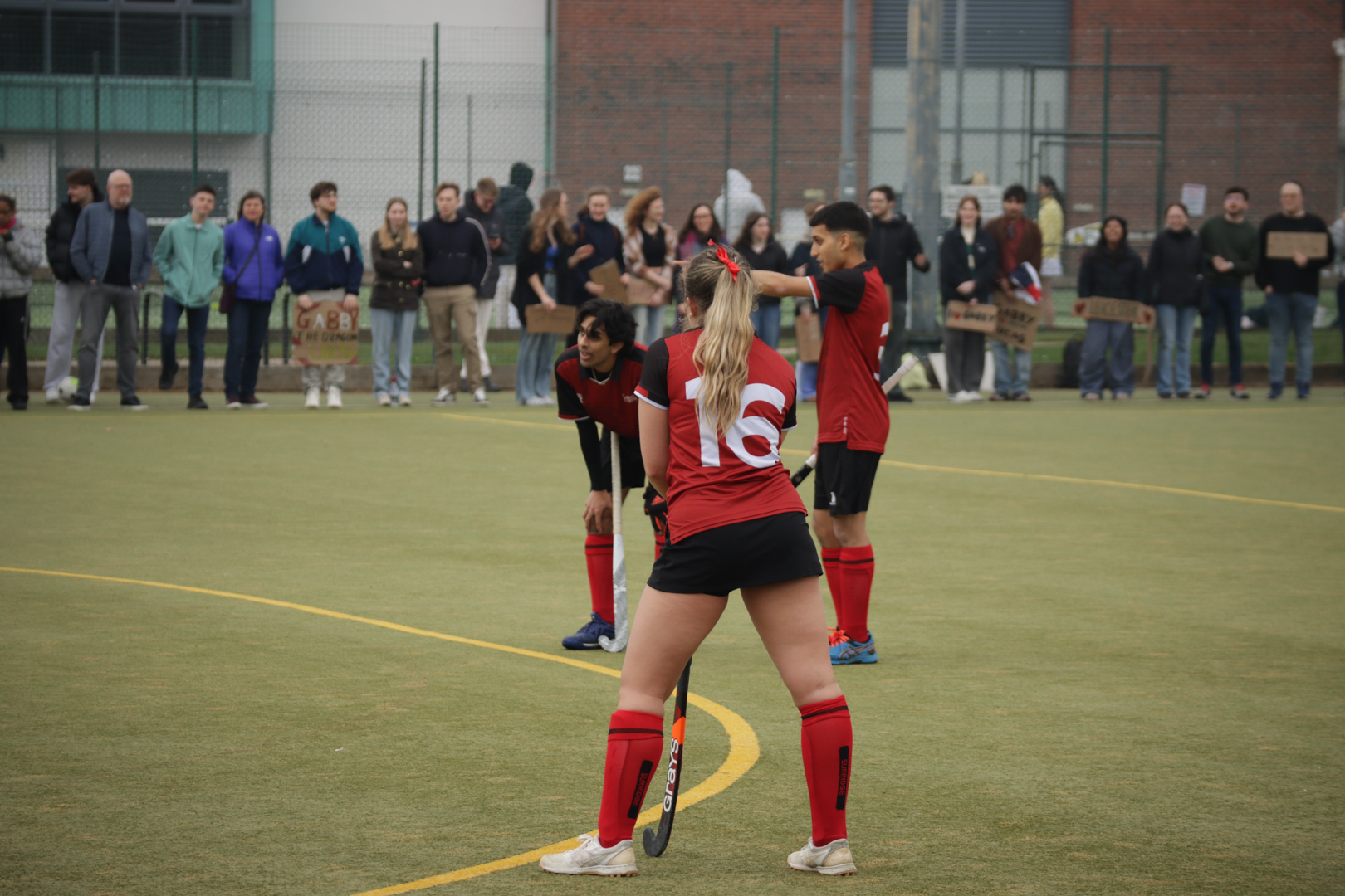 Hockey players in red and black uniforms prepare to start play on a turf field with spectators in the background.