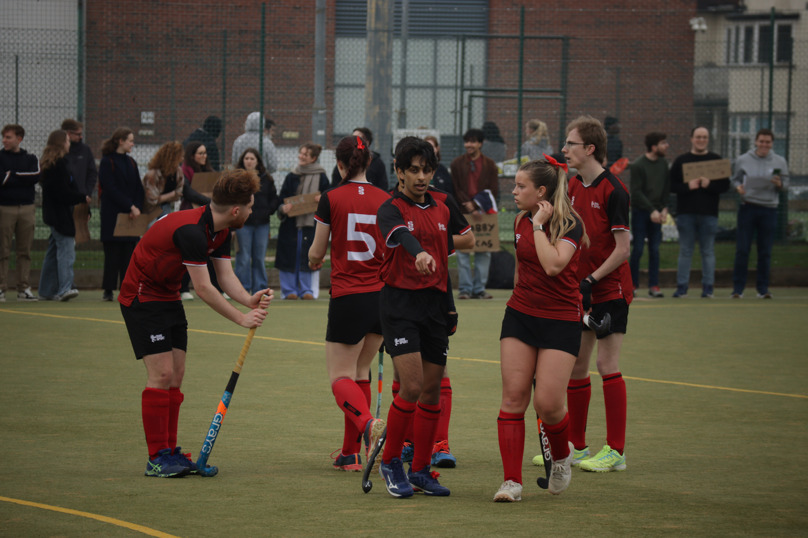 Hockey players in red and black uniforms standing together on a field. One player giving instructions.