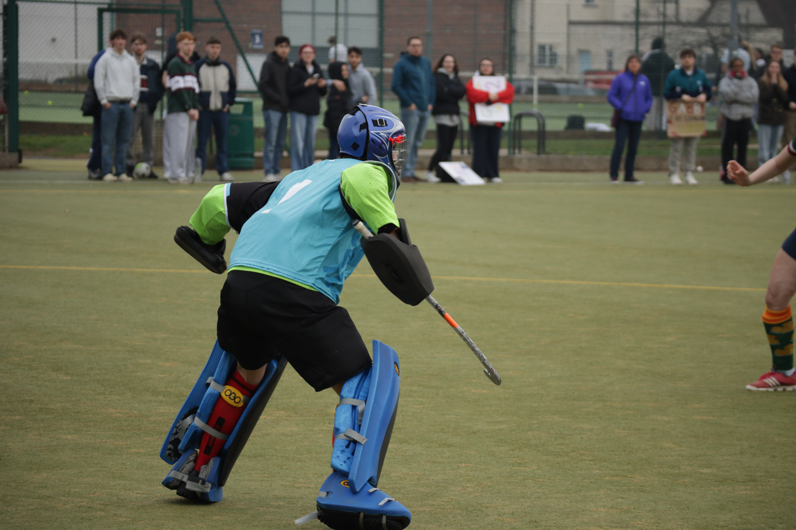 Hockey goalie in protective gear faces an approaching player on a turf field, with spectators in the background.