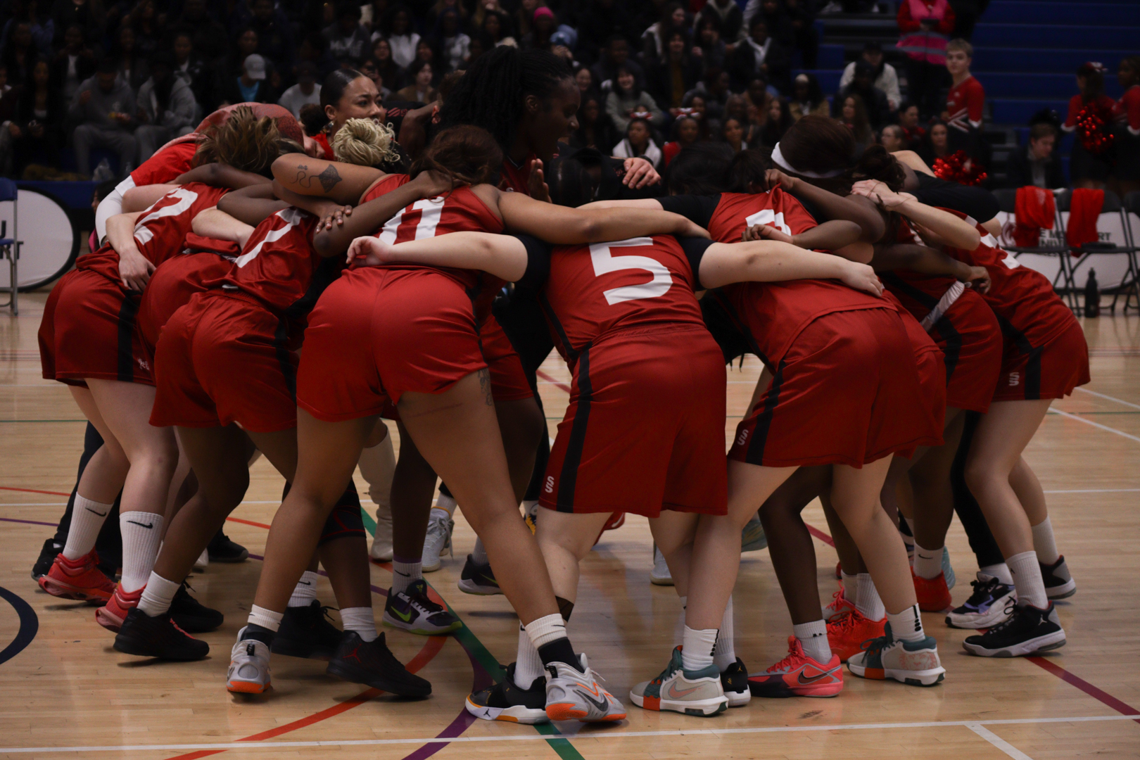 Basketball team in red kits huddling together on a court before a game, with spectators in the background.