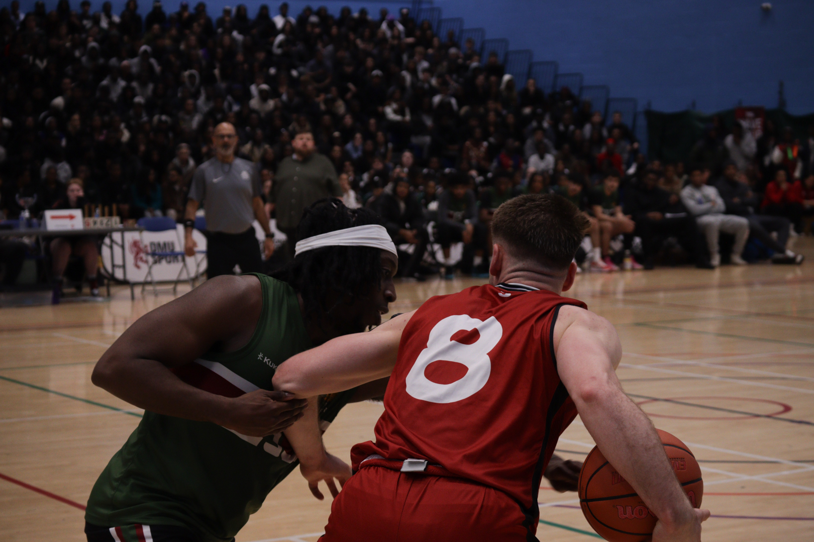 Two basketball players compete for the ball during a game, with a crowd watching in the background.