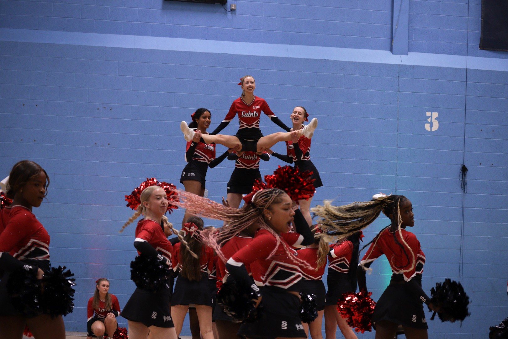 Cheerleaders in red and black uniforms perform a group stunt indoors, lifting one member above the rest.