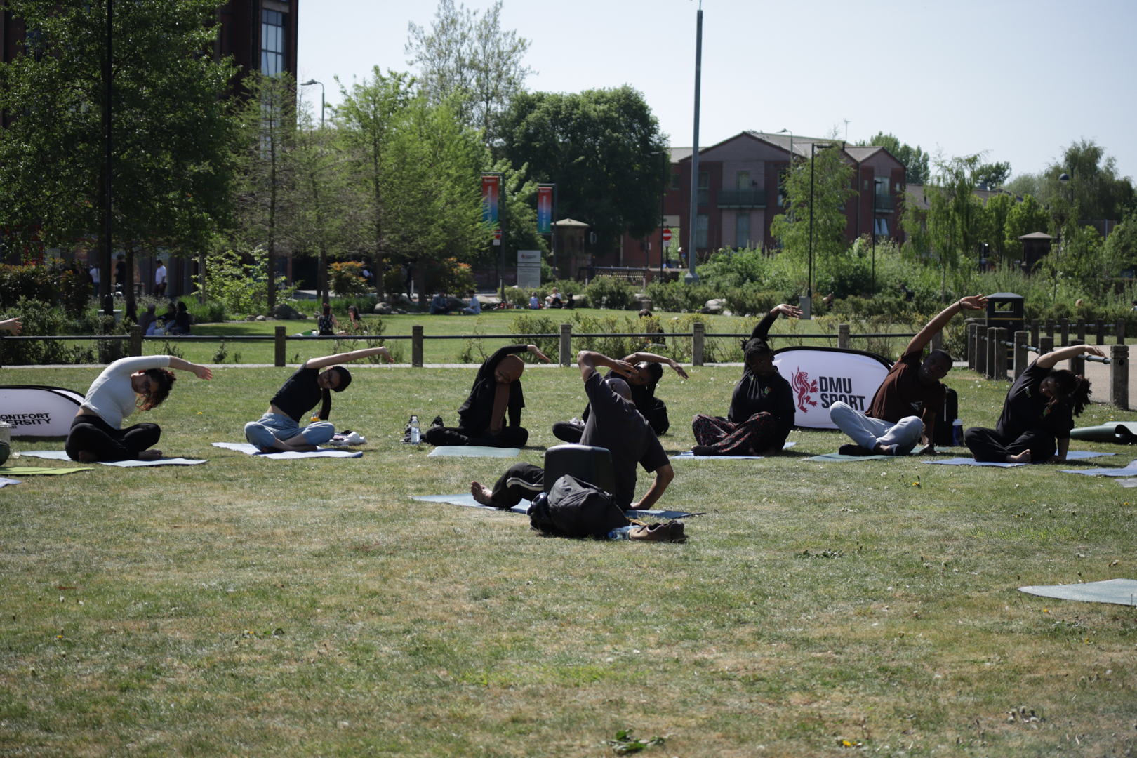 People practicing yoga outdoors on a grassy field, each on a mat, with trees and buildings in the background.
