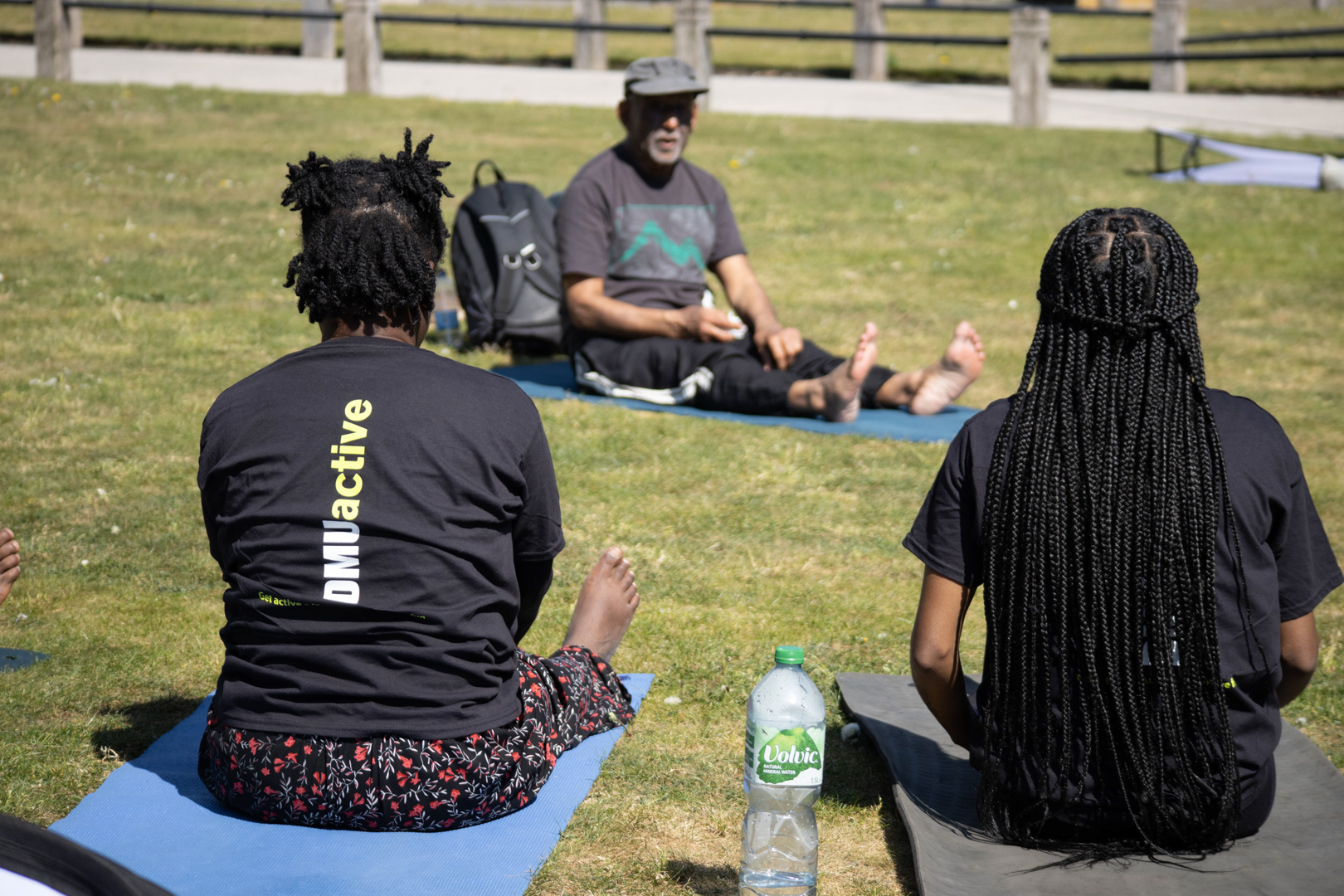 Three people sit on mats on grass, with one person facing the others and leading an outdoor session.