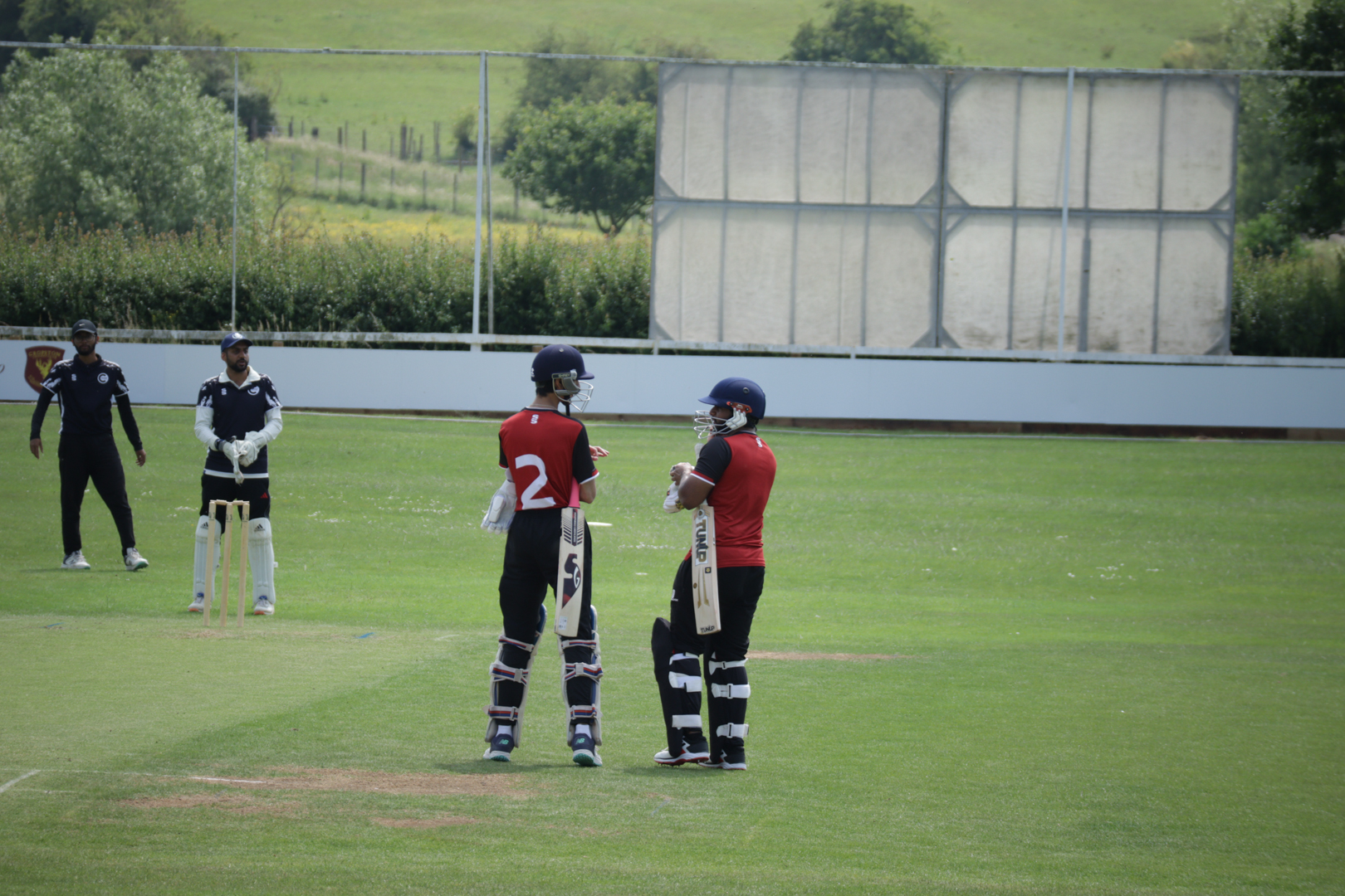 Two cricket players in red shirts stand on a field, with other players in the background.