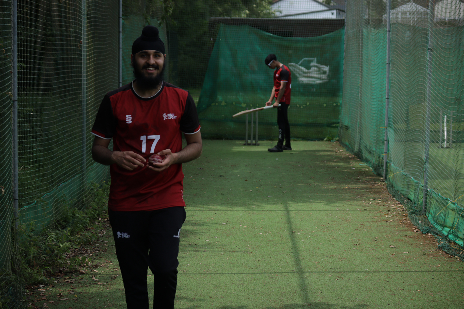 Two people in a cricket practice net, one holding a ball and the other preparing to bat.