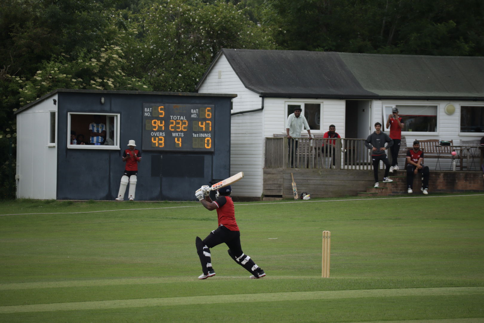 Cricket batsman playing shot on a grassy field with scoreboard and small buildings in the background.