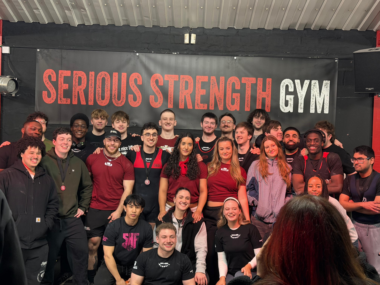 Large group of people posing and smiling inside a gym with a "Serious Strength Gym" sign on the wall.