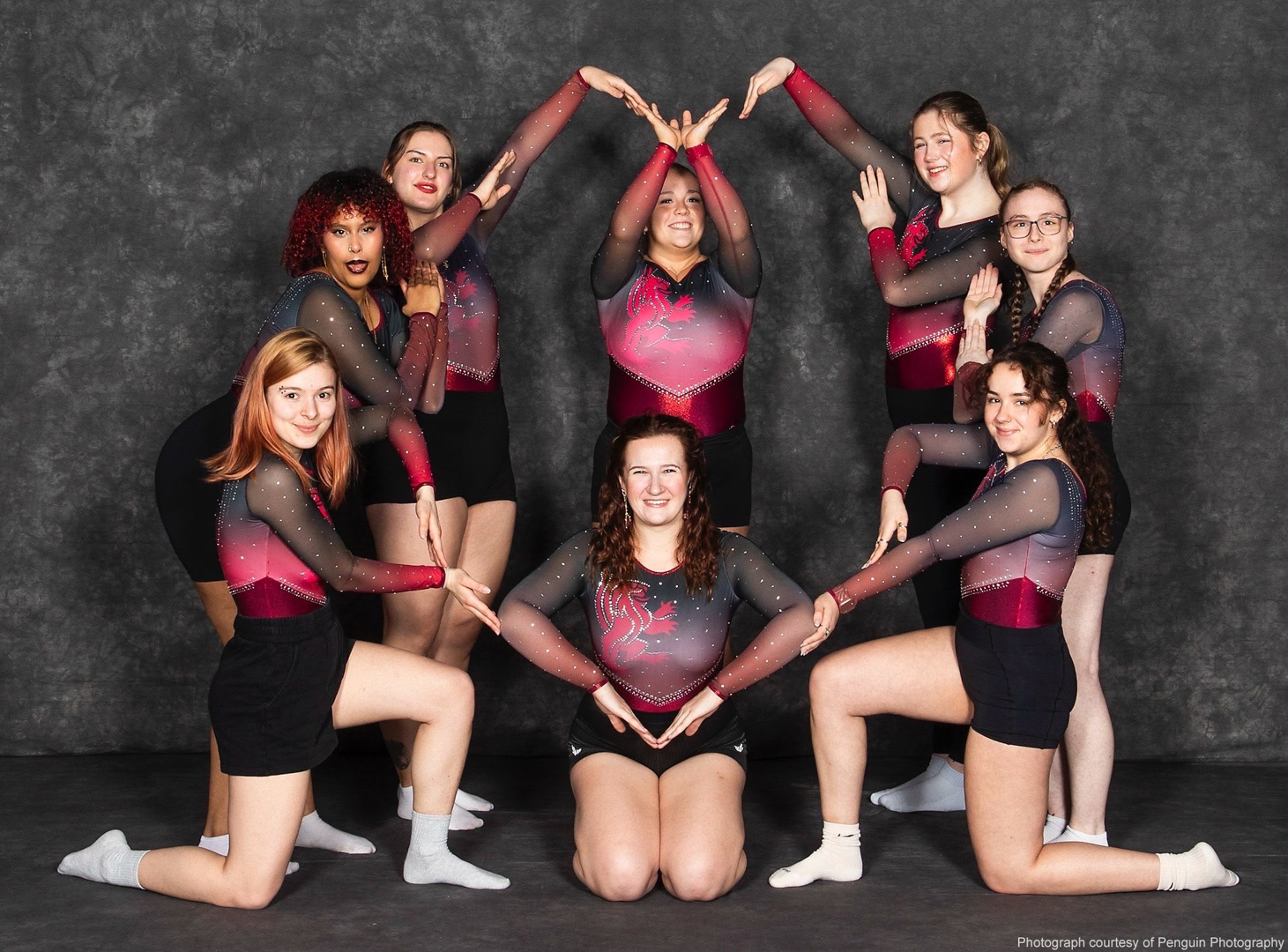 Eight students in matching uniforms pose together, forming a heart shape with their arms against a dark background.