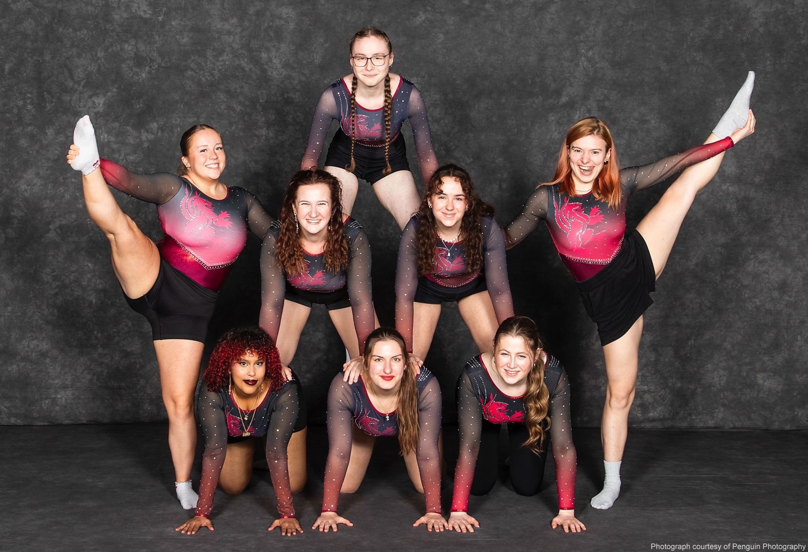 Seven students pose in a pyramid formation, with two holding high leg extensions on either side.
