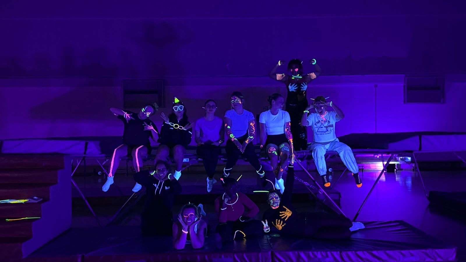 A group of people sitting together on a trampoline in a dark room with glow sticks and UV lights.