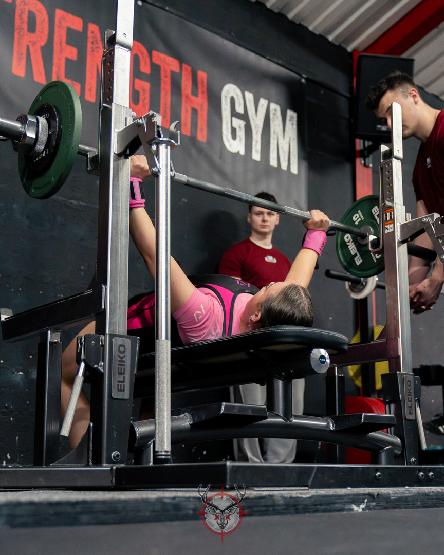 Woman performing bench press with barbell in a gym while two people assist on either side.
