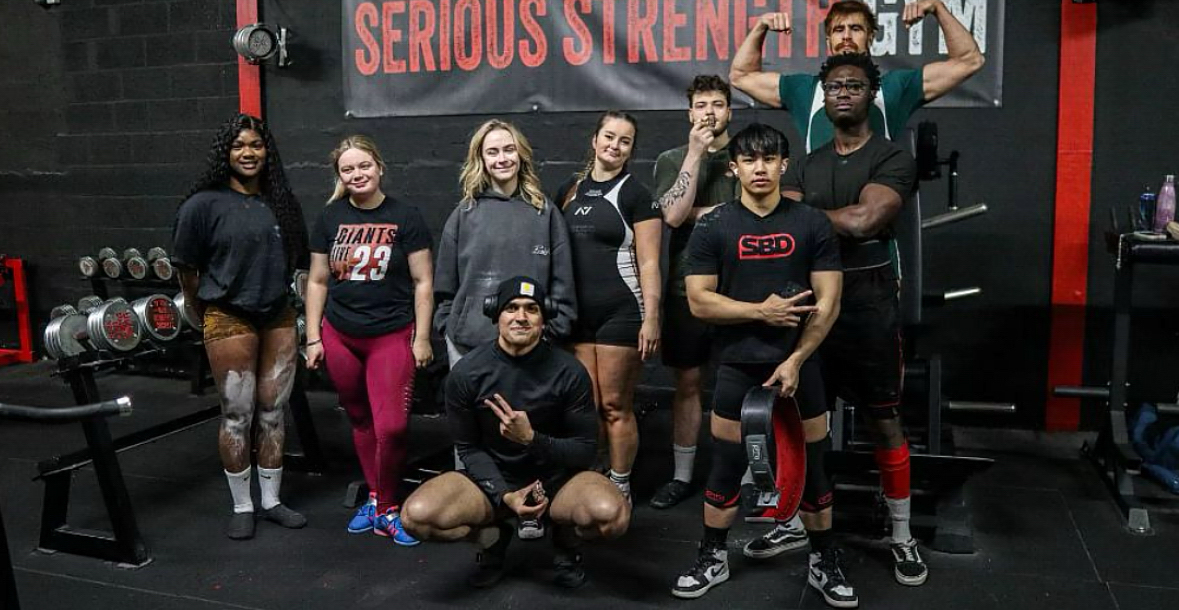 People in athletic wear posing together in a gym with weightlifting equipment and a banner in the background.