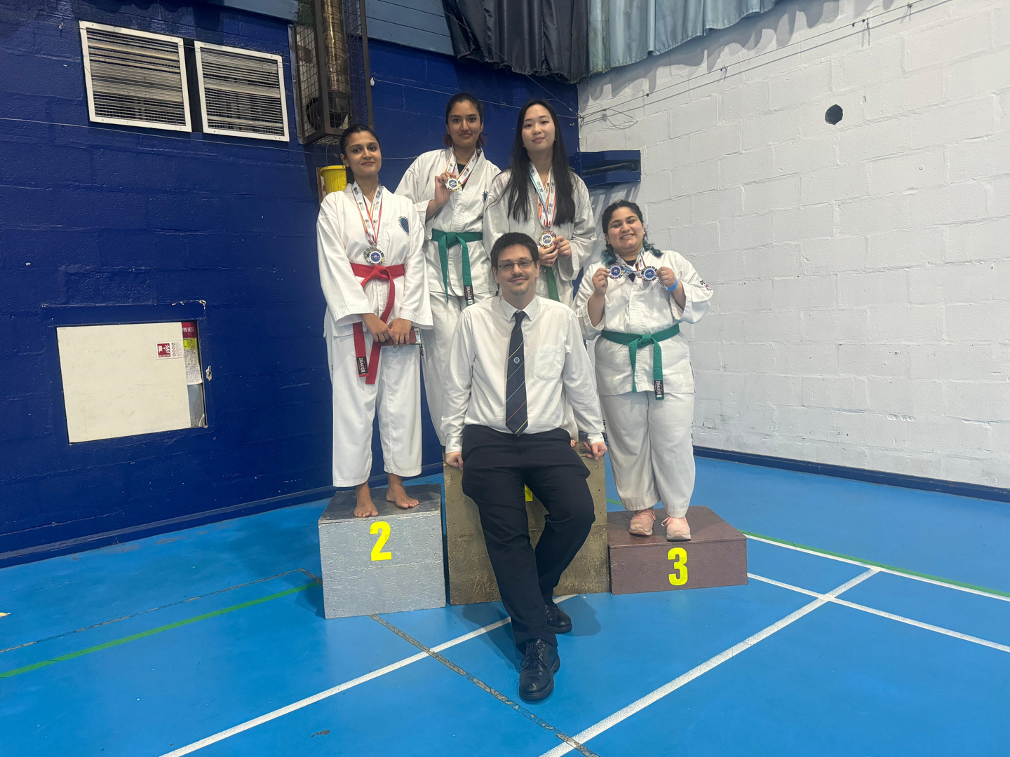 Four martial artists in uniforms and belts stand on a winners' podium with medals in a gymnasium.