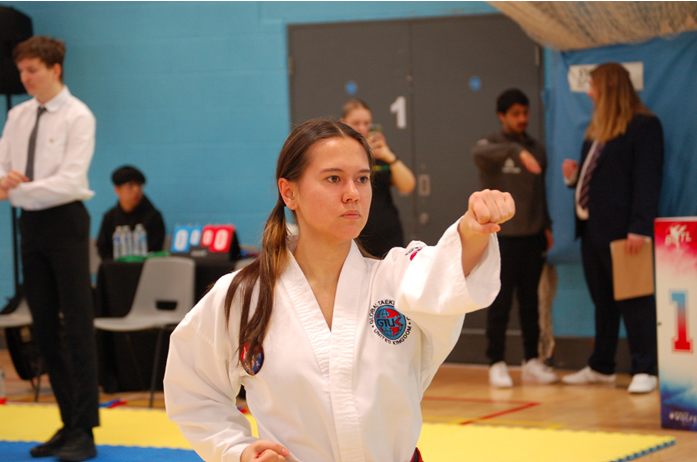 Martial artist in a white uniform performing a punch during a competition, with a referee in the background.