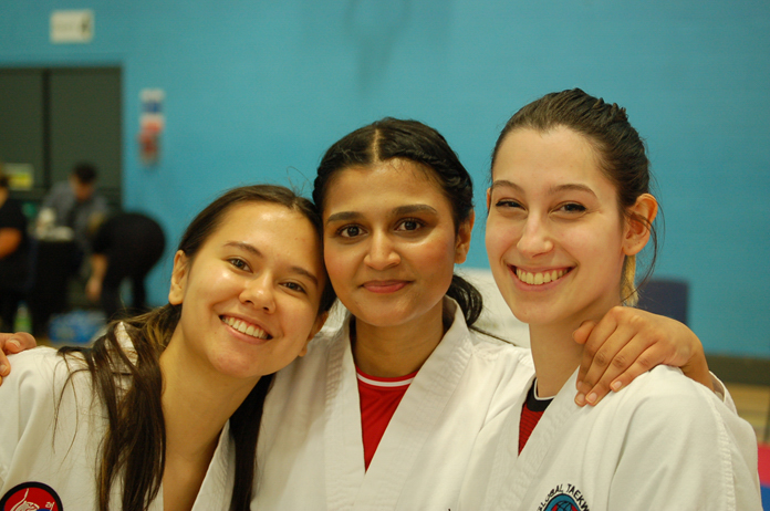 Three women in martial arts uniforms smiling and posing together in an indoor gym setting.