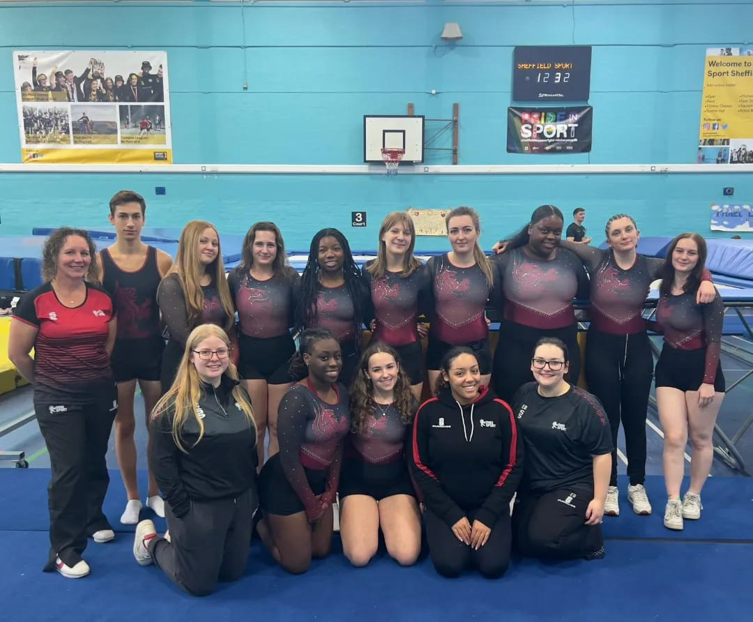A group of athletes posing together in a gymnasium, with trampolines behind them.