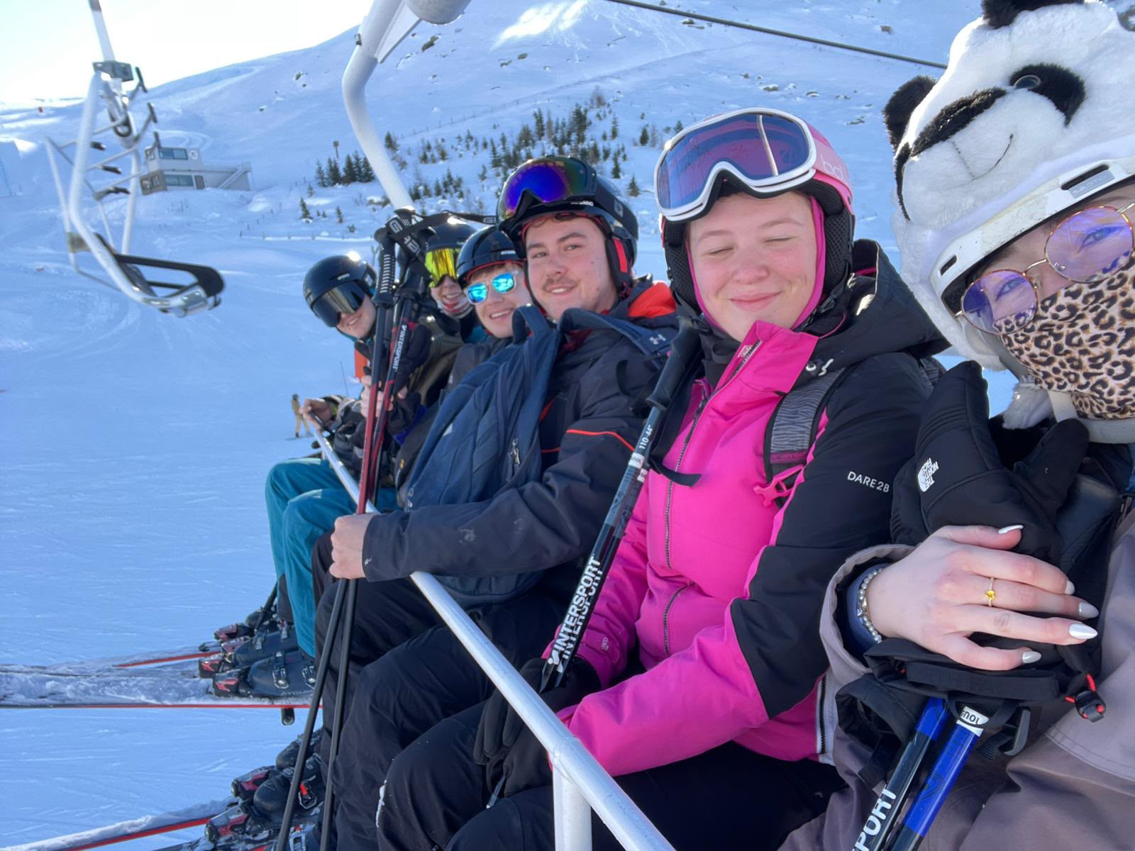 Four people in winter gear sitting on a ski lift with snowy mountains in the background.