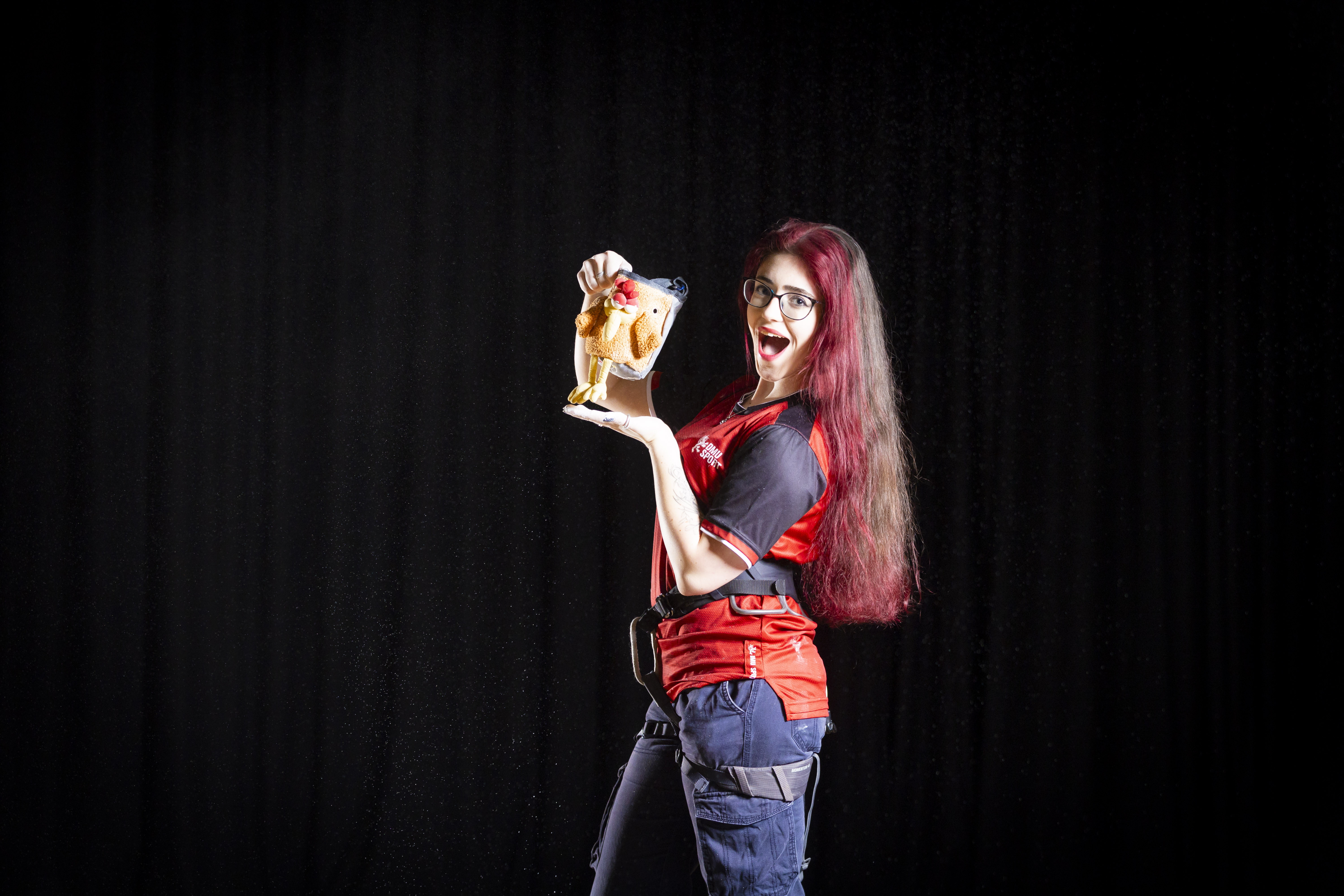 Woman with long hair and red shirt holding a chalk bag, smiling against a dark background.