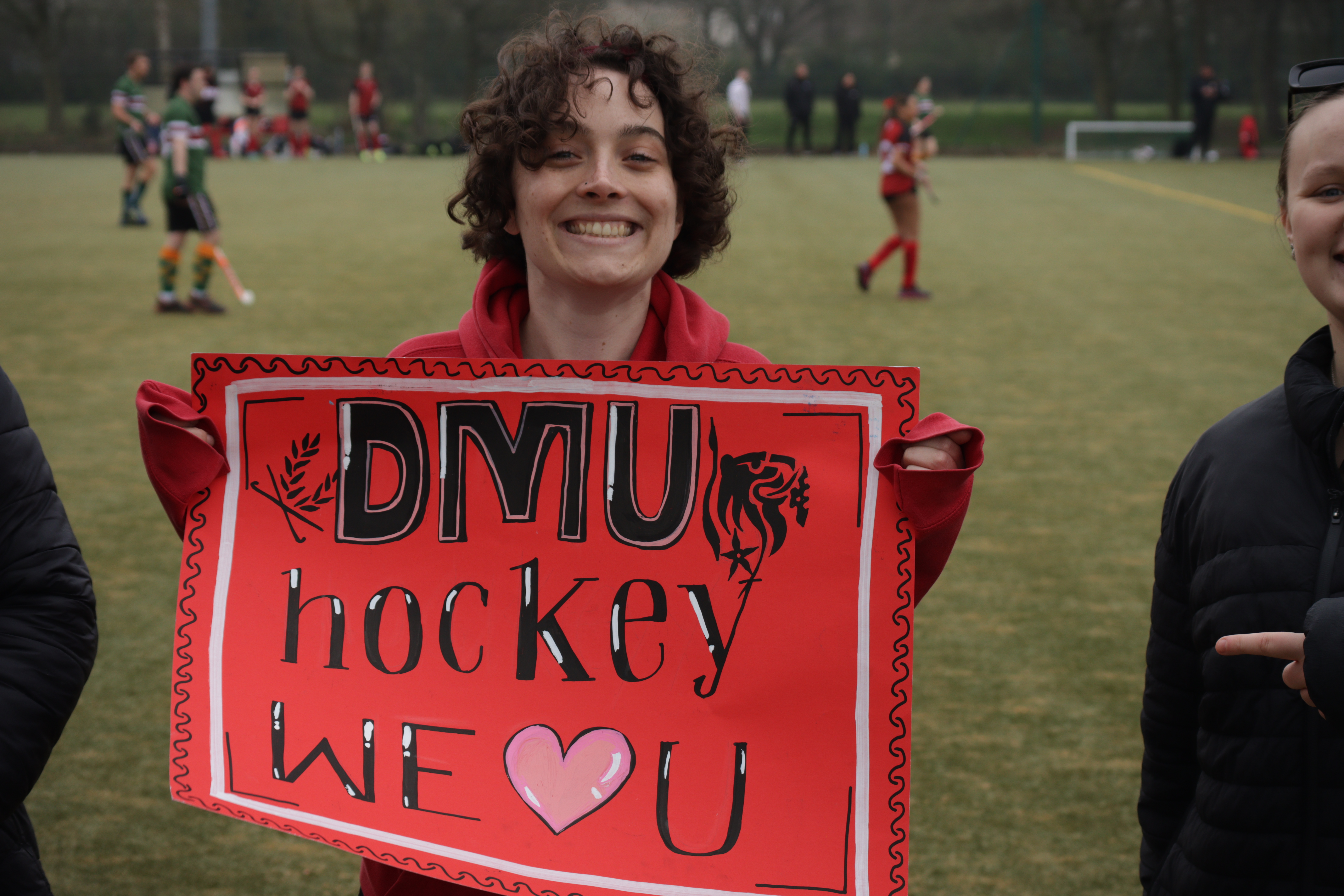 Person smiling and holding a red sign that says "DMU Hockey We Love U" on a sports field with players in the background.