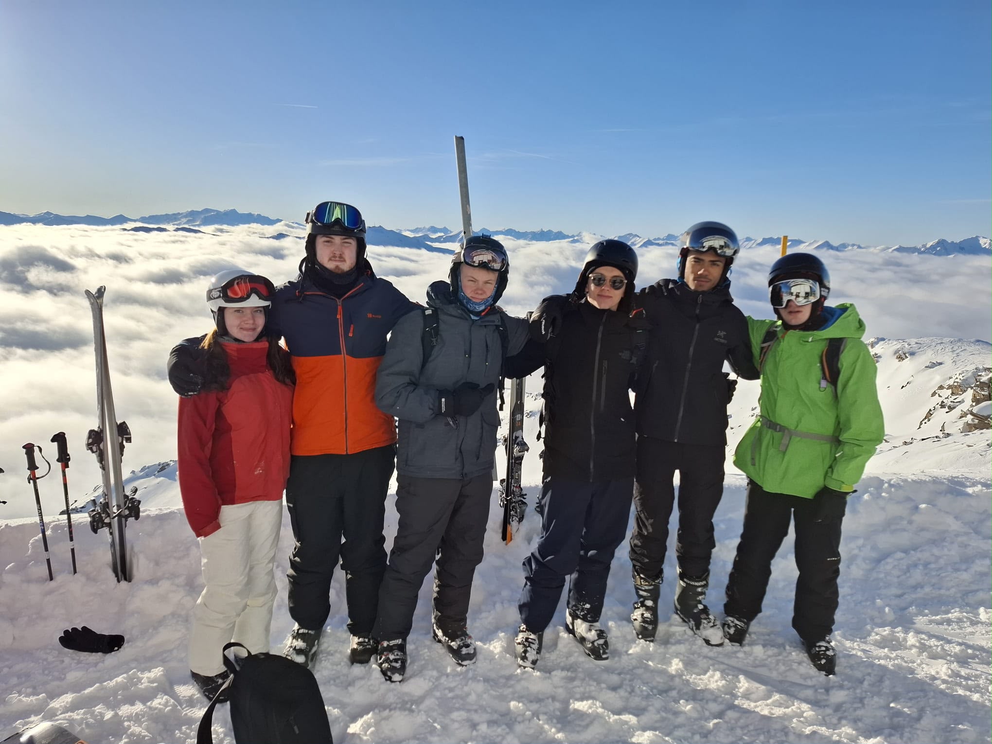 Six people standing together on a snowy mountain with ski poles and snow-covered hills in the background.